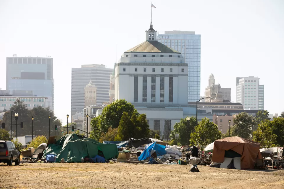 Unhoused encampment on dry lawn with Art Deco building in background in Oakland, California.